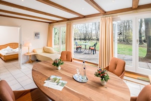 Habitación de hotel con una mesa de comedor de madera, sillas, una cama, un sofá y vistas a un jardín a través de amplios ventanales.