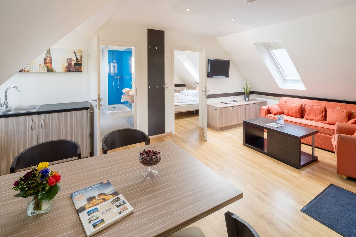 Una habitación de hotel moderna con zona de comedor, kitchenette y sala de estar con sofá y televisor.
