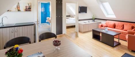 A modern hotel room with a dining area, kitchenette, and a living space with a sofa and a TV.