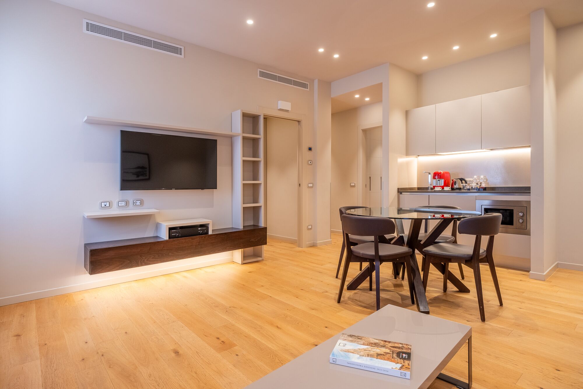 A modern living room with a flat-screen TV, a coffee table, and a dining area with chairs.