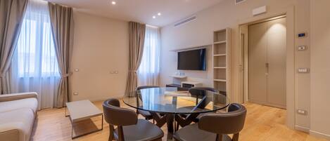 A modern hotel room with a glass dining table, four chairs, a sofa, a TV, a wardrobe, and a small bedside table.