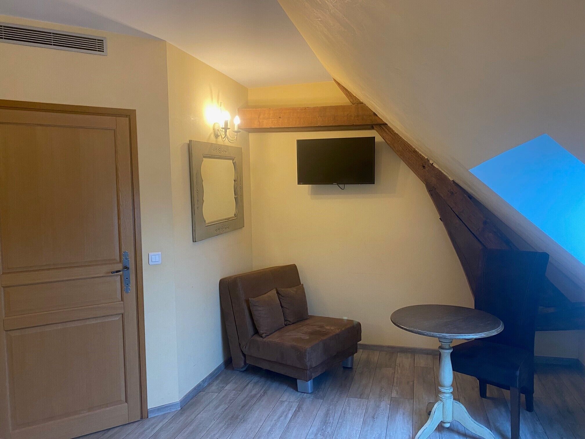 Ein Zimmer mit Holztür, einem kleinen braunen Sofa, einem runden Tisch und einem flachbildfernseher an der Wand.