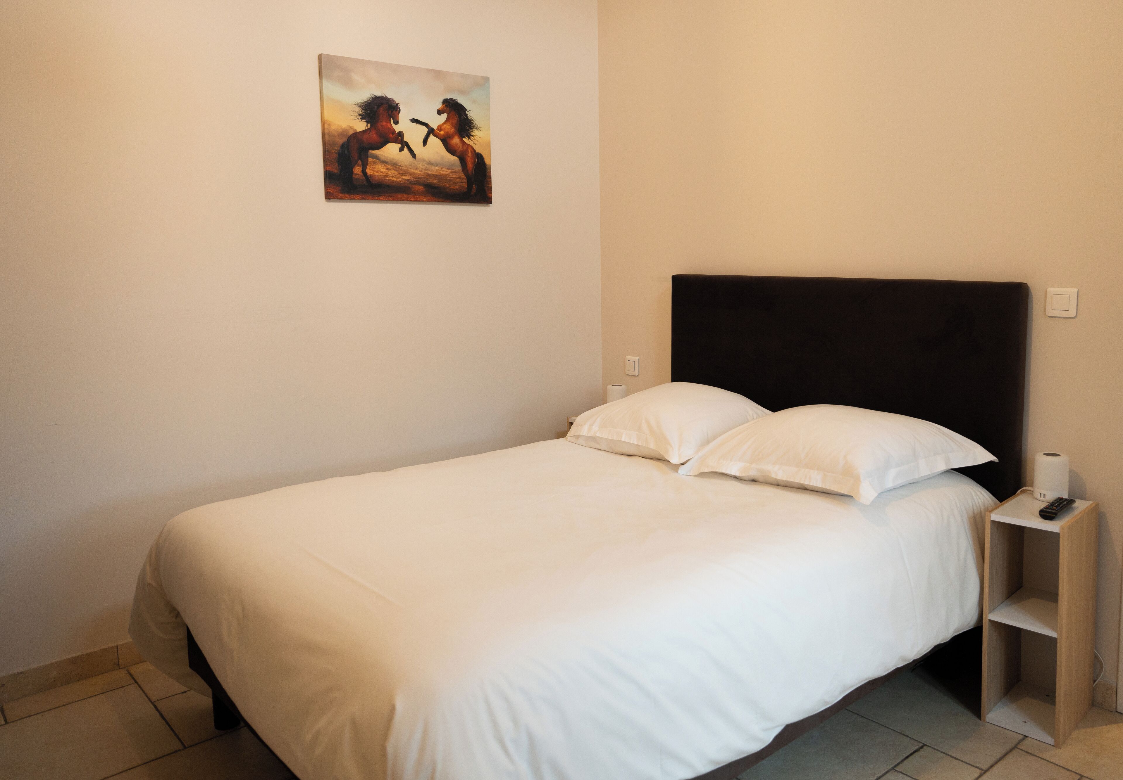 A neatly made bed with white linens, a headboard, and a small bedside table with a remote control and a roll of toilet paper.