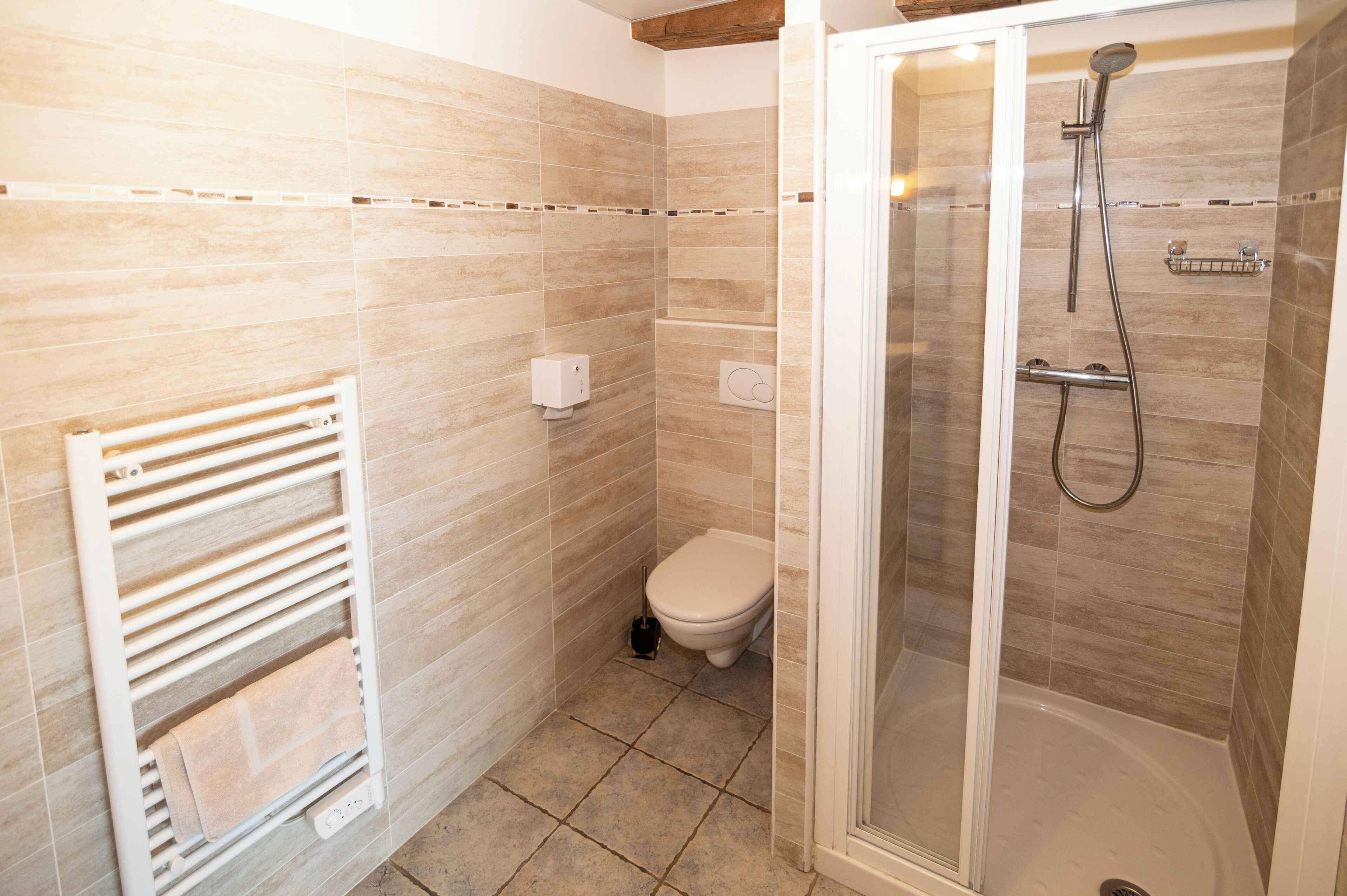 A bathroom with beige tiles, a white toilet, a glass-enclosed shower, and a towel rack.