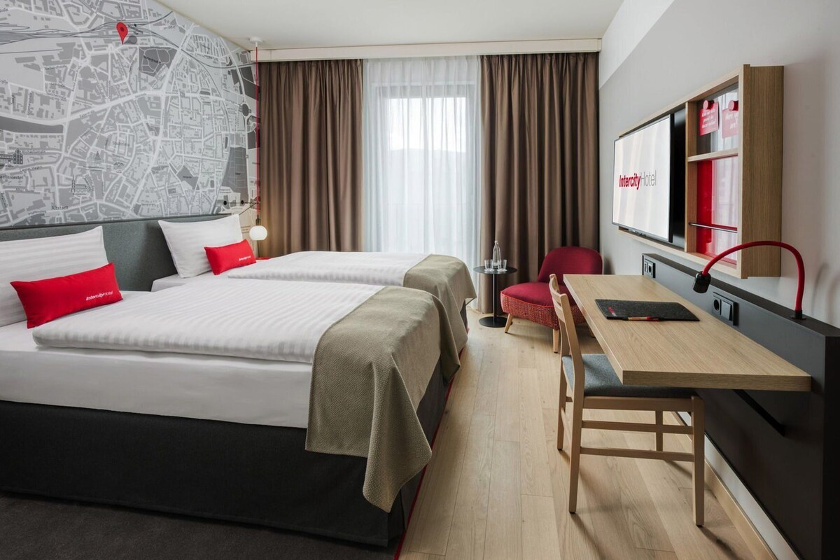 Habitación de hotel con dos camas, un escritorio, una silla, un mapa en la pared y un cartel que dice 'Intercity hotel'.