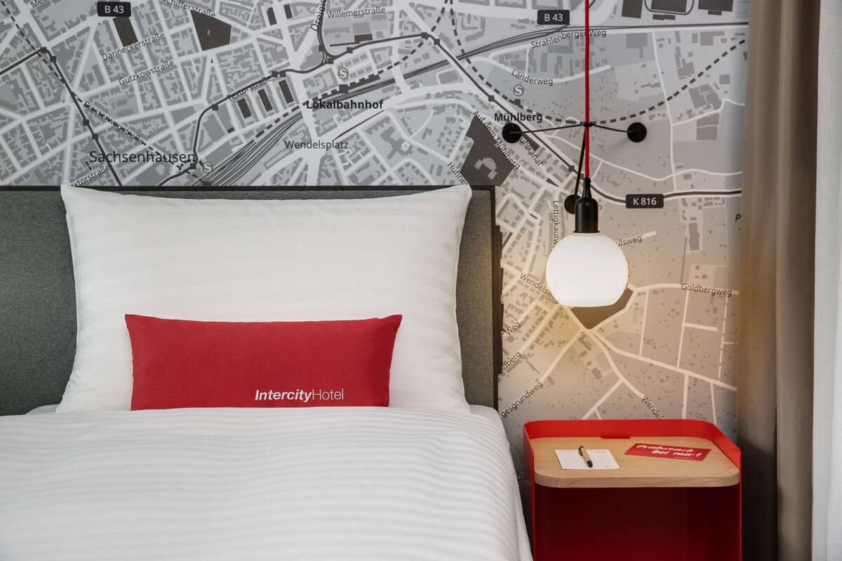 Une chambre d’hôtel avec un lit, une table de chevet, une carte accrochée au mur et un oreiller rouge.