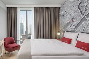 Un cuarto de hotel con una cama grande, almohadas rojas y blancas, un mapa en la pared, una silla y vistas de la ciudad por la ventana.