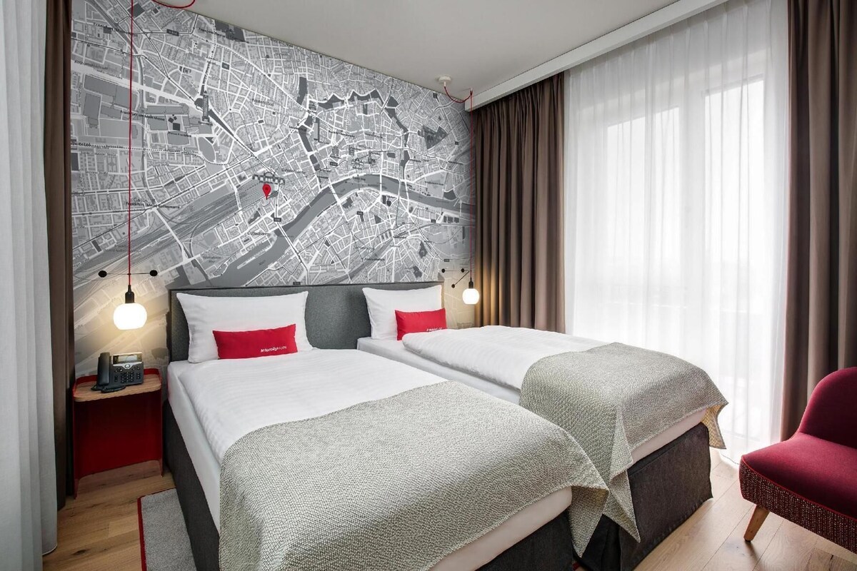 Habitación de hotel con un mapa de la ciudad en la pared, dos camas con ropa de cama roja y blanca, una silla roja y una mesita de noche con un teléfono.