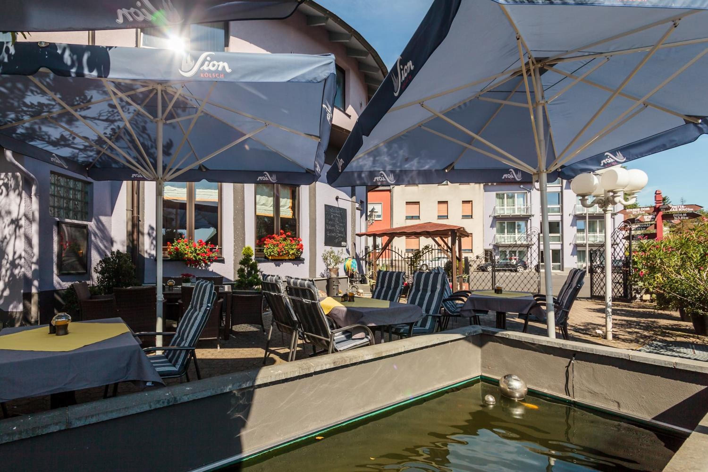 An outdoor seating area with umbrellas, tables, and chairs, a small pool, and buildings in the background.