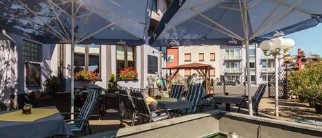 An outdoor seating area with umbrellas, tables, and chairs, a small pool, and buildings in the background.