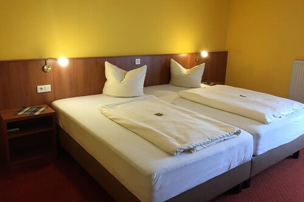 A double room with two beds, each with a pillow and a blanket. There is a nightstand with a lamp and a book. The walls are yellow, and there is a wooden headboard.