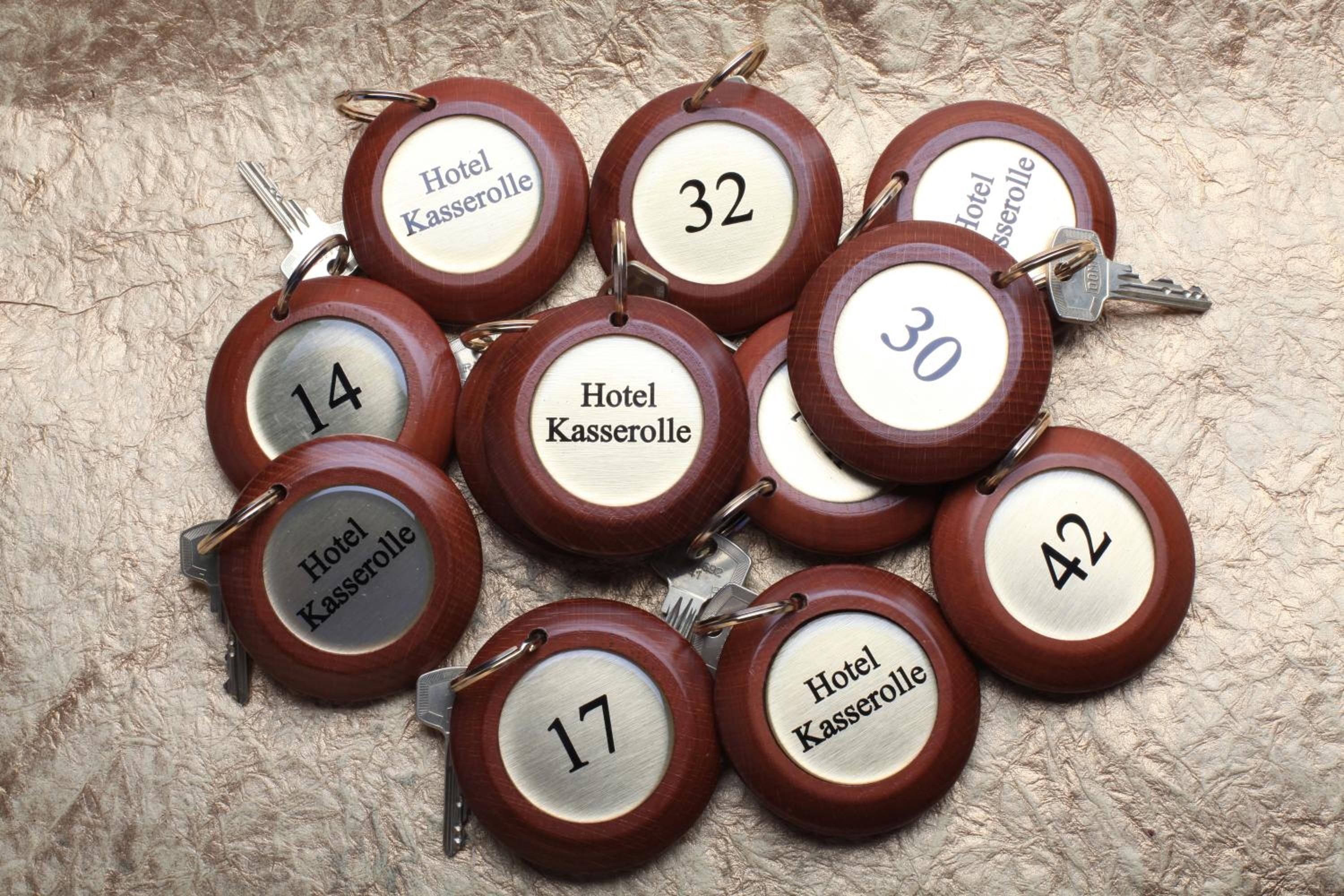Eine Auswahl aus Holz-Schlüsselanhängern mit Zimmernummern und dem Schriftzug 'Hotel Kasserolle' auf einer strukturierten Oberfläche.
