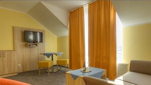 A hotel room with a television mounted on the wall, a small table with a tablecloth, a sofa, and a window with curtains. - Hotel Restaurant Kasserolle (Siegburg)