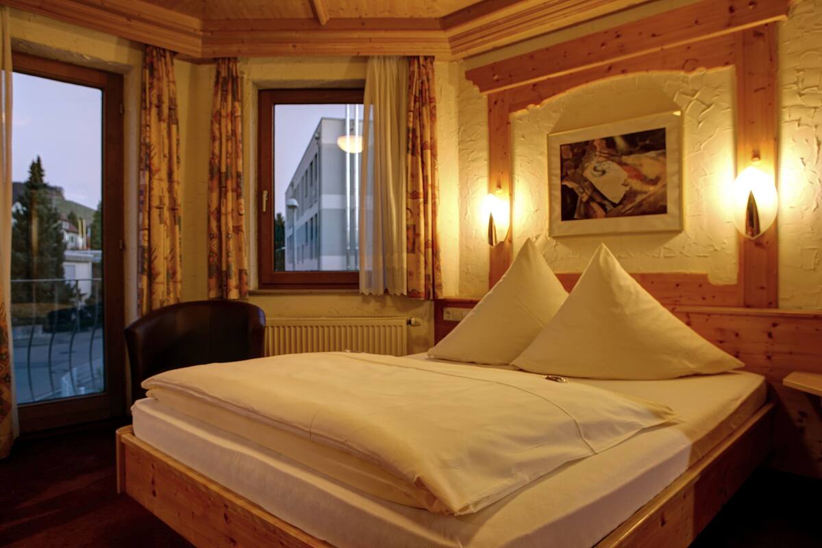A wooden-framed room with a bed, a chair, a window with curtains, and a wall-mounted light fixture.