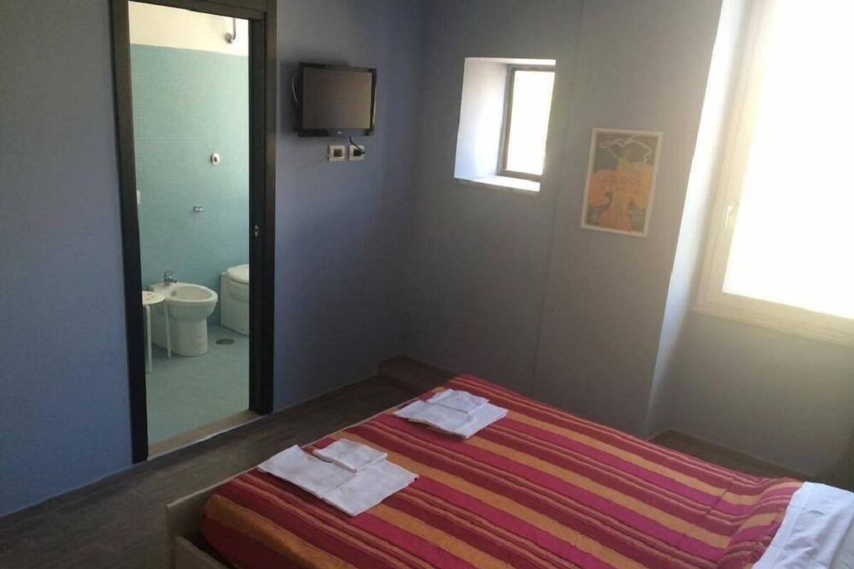 Ein kleines Zimmer mit einem Einzelbett, einer gestreiften Bettdecke, einem Fernseher an der Wand, einem kleinen Fenster und einem durch eine offene Tür einsehbarer Badezimmer.