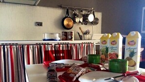 Un coin cuisine avec un plateau rouge contenant une assiette, des ustensiles et des aliments. On y voit des casseroles et des poêles suspendues, un plan de travail blanc et une étagère avec divers articles de cuisine.
