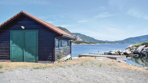 Une cabane en bois avec une porte verte, située au bord d’un lac paisible aux rives rocheuses, avec des montagnes en arrière-plan.