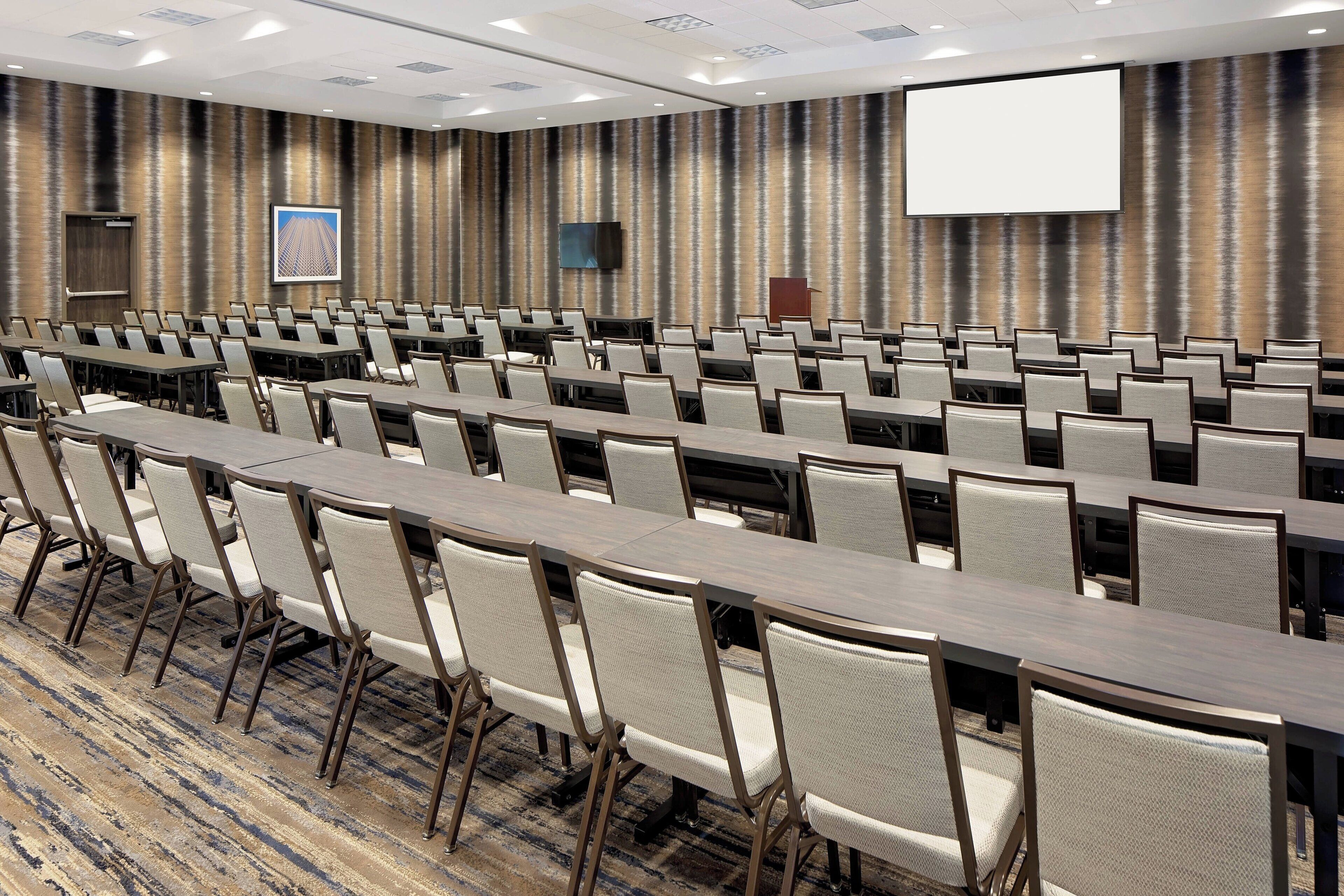 A conference room with rows of tables and chairs, a large screen, and a patterned wall.