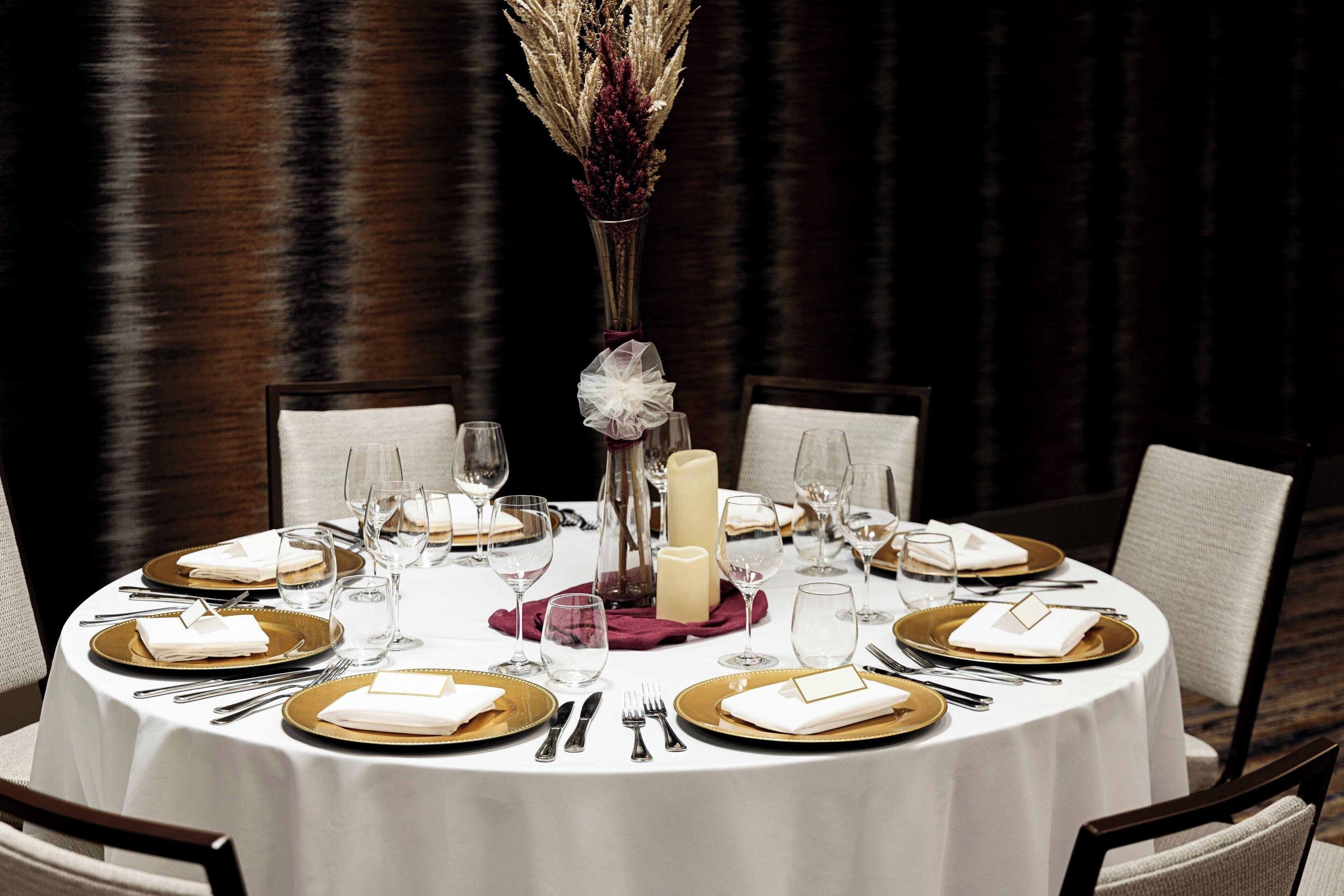 A round dining table set with gold-rimmed plates, white napkins, and silverware. A tall vase with dried flowers is centered on the table.