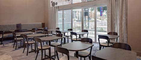 A hotel dining area with round tables and chairs, a patterned carpet, and large windows offering a view of the street.