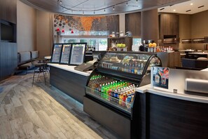 Interior moderno de una cafetería con una barra que exhibe diversas bebidas, una zona de estar con mesas y sillas, y un amplio ventanal que proporciona luz natural.