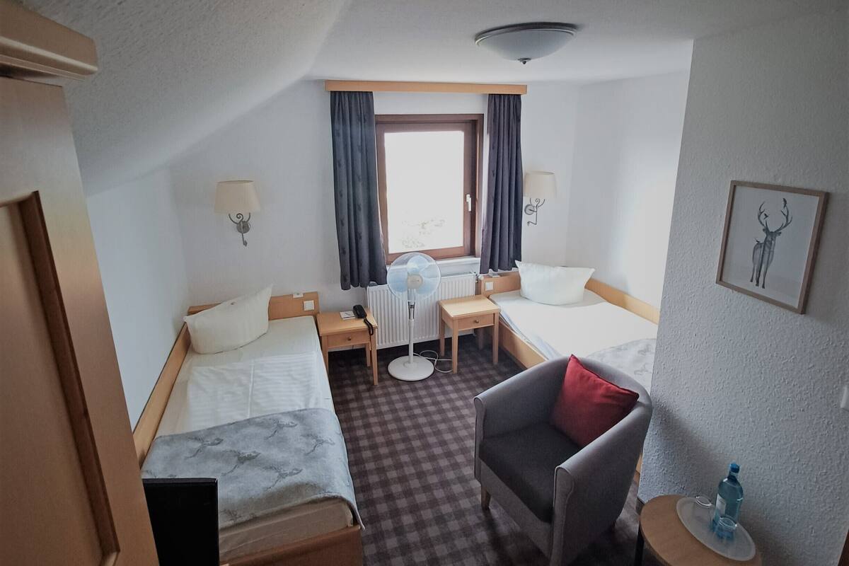 Une chambre d’hôtel avec deux lits, un bureau, une chaise, une petite table, un radiateur, une fenêtre avec des rideaux et un ventilateur de plafond.