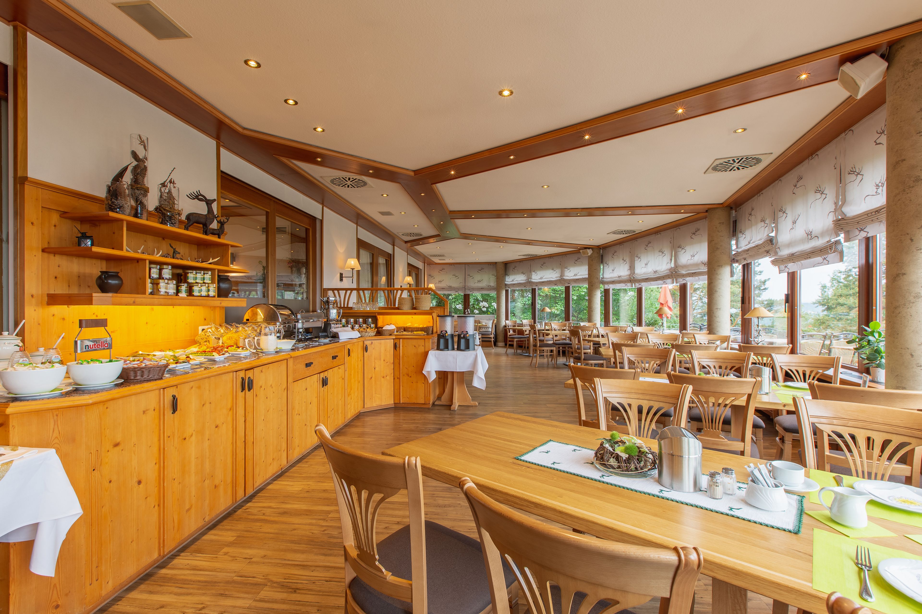 A spacious dining area with wooden tables and chairs, a buffet counter with various food items, and large windows providing natural light.