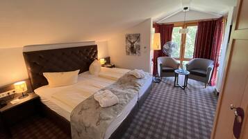 A hotel room with a large bed, a nightstand with a lamp, a chair, a small table, and a window with curtains.