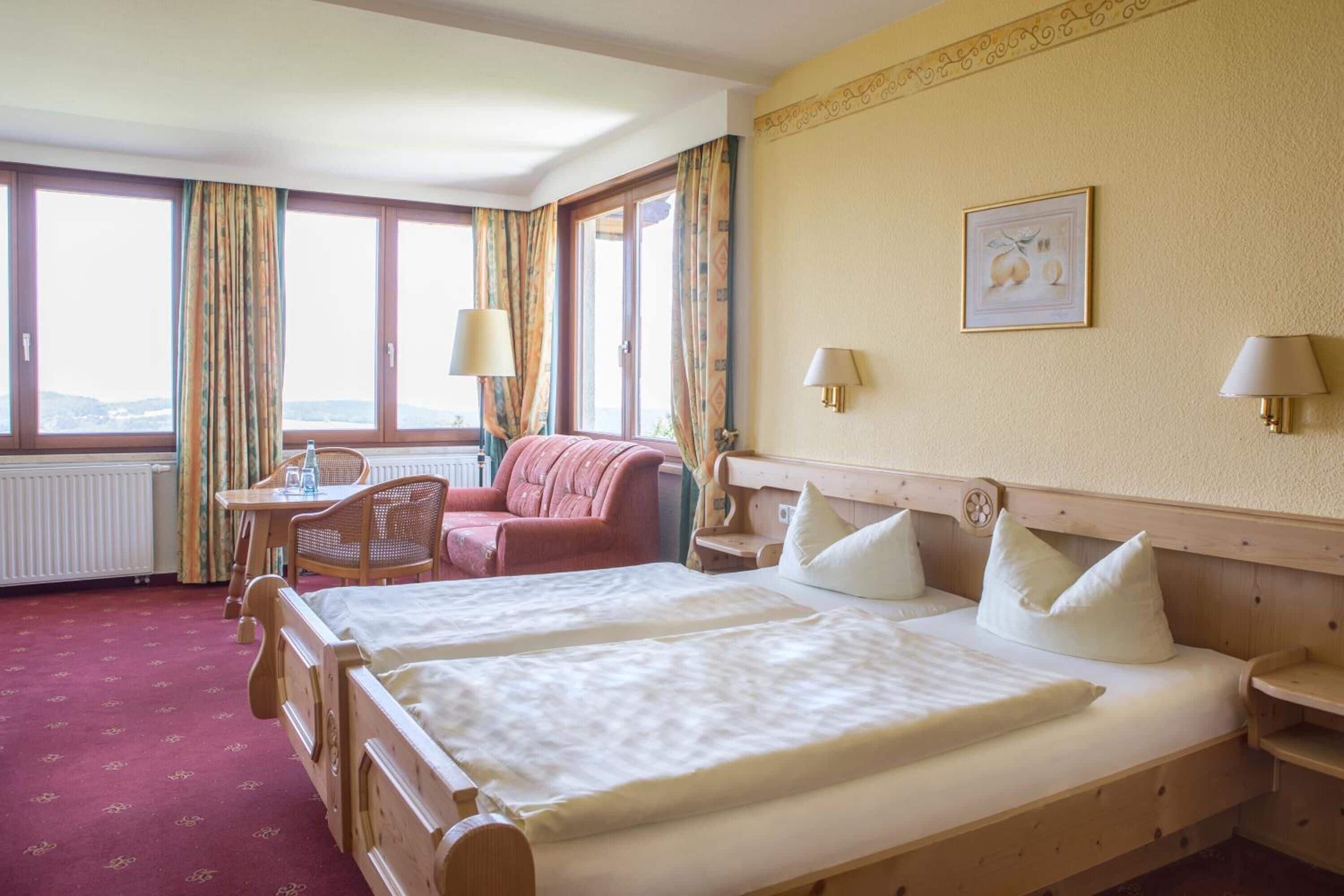 Une chambre d’hôtel avec deux lits, un coin salon avec une chaise et une table, une vue dégagée grâce à de grandes fenêtres et une lampe fixée au mur.