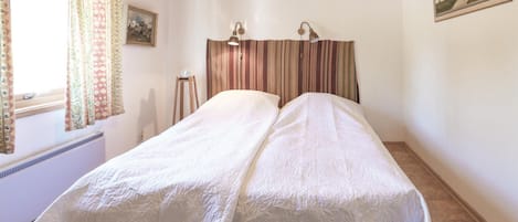 Een tweepersoonsbed met een witte sprei, een houten hoofdbord, twee wandlampen en een raam met gordijnen.