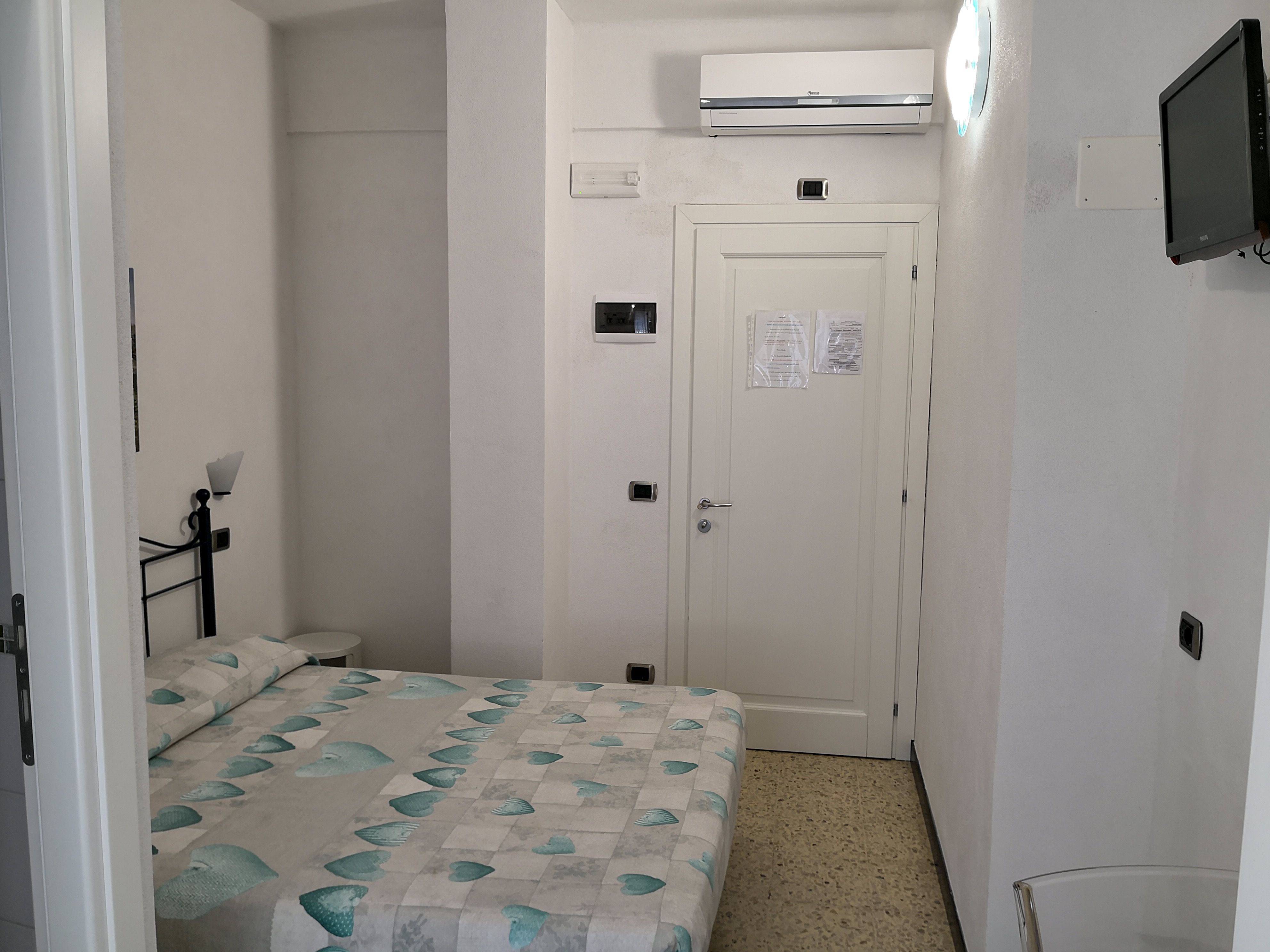 Une petite chambre d’hôtel étroite, comprenant un lit, une table de chevet, un téléviseur fixé au mur et un appareil de climatisation.