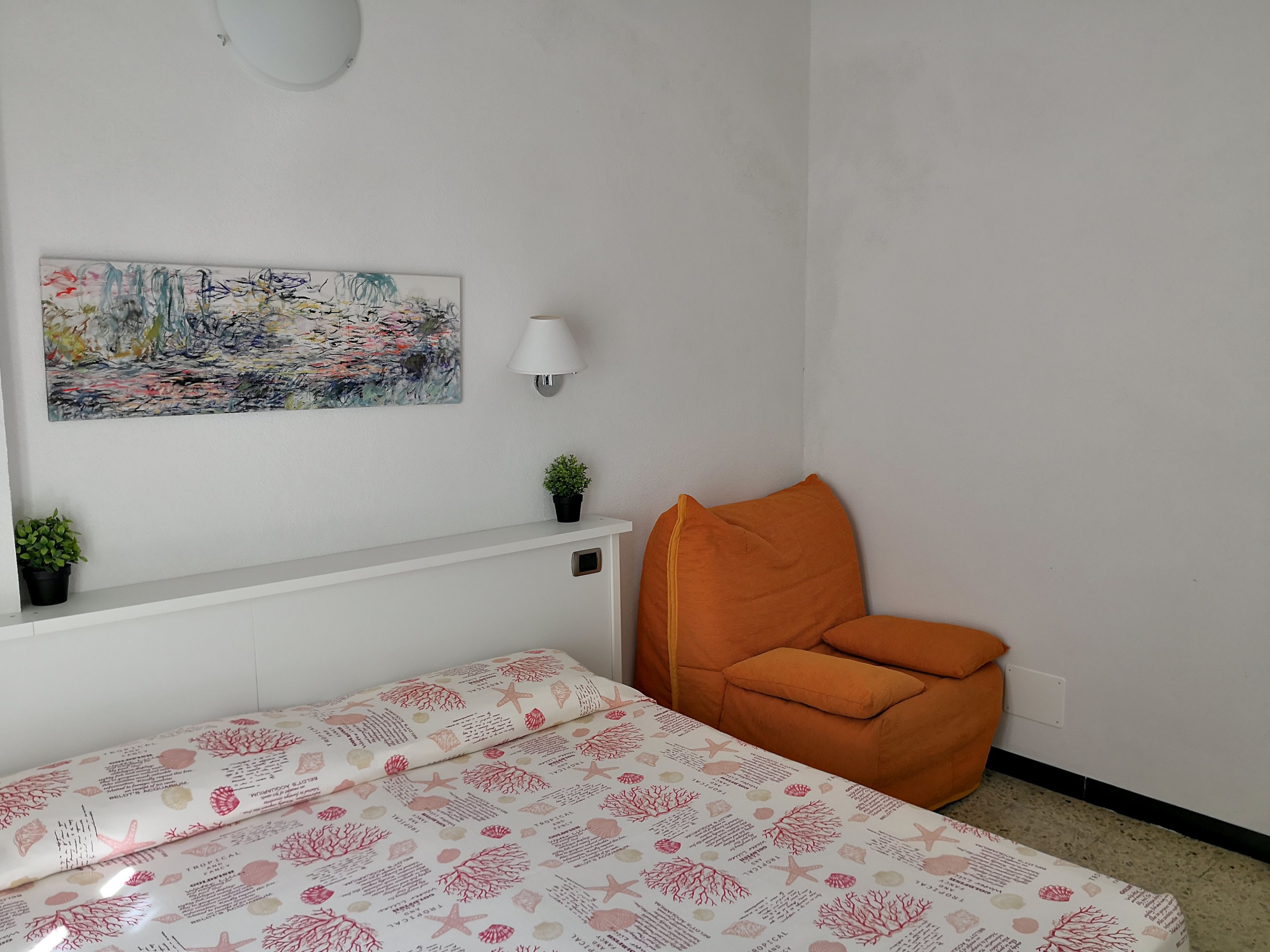 Et værelse med en seng, en orange stol, et lille bord med en plante, en væglampe og et farverigt abstrakt maleri.
