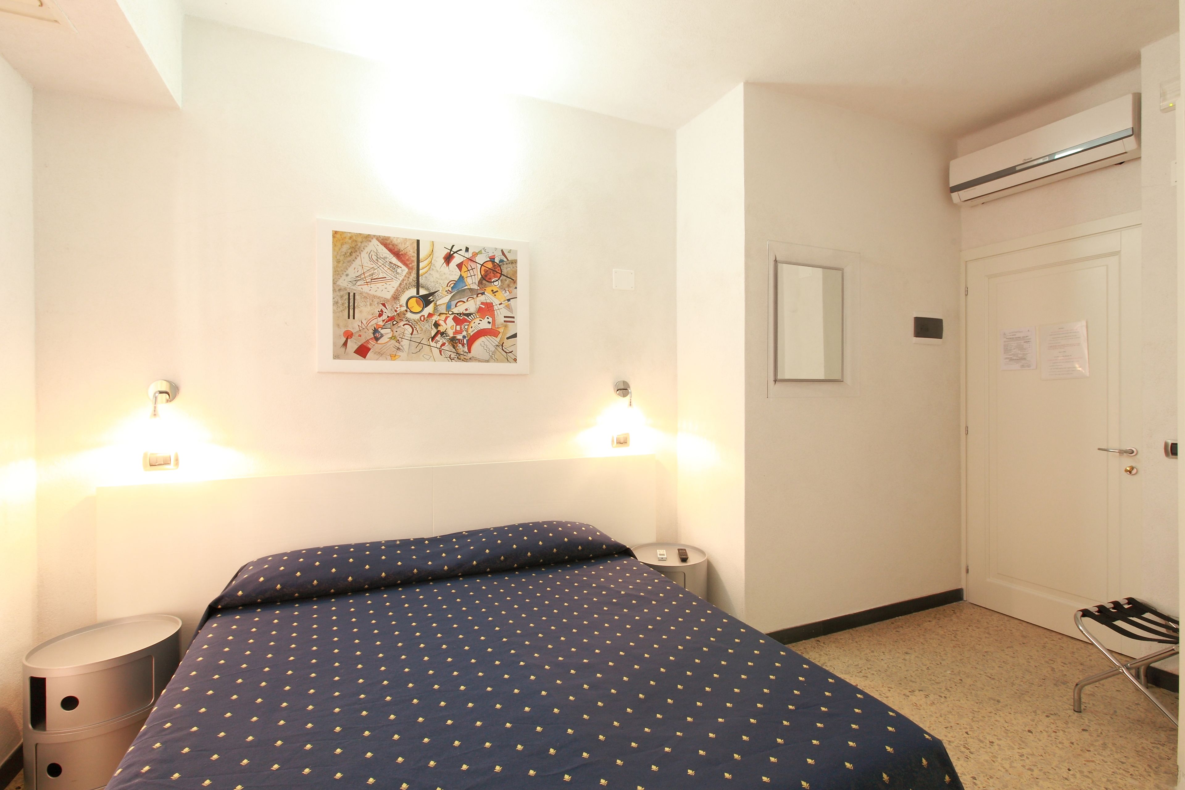 A hotel room with a bed, bedside lamps, a painting on the wall, a door, and an air conditioning unit.