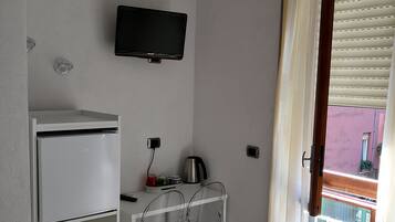 Habitación de hotel pequeña con una cama, un escritorio con silla, un televisor montado en la pared y una ventana con persianas.