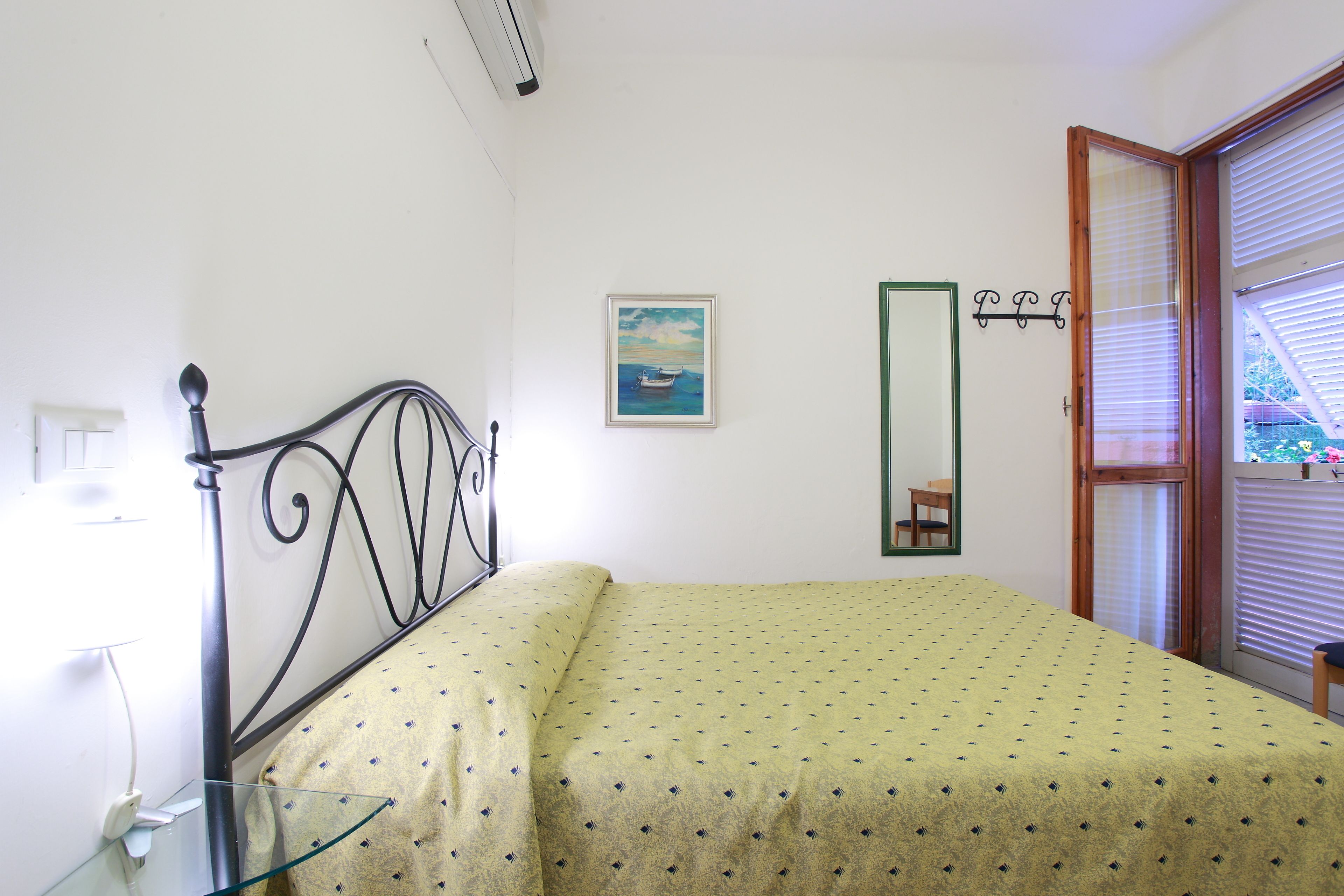 A bedroom with a bed, a bedside table, a mirror, a window with blinds, and a painting on the wall.