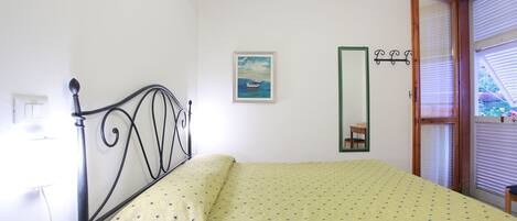 A bedroom with a bed, a bedside table, a mirror, a window with blinds, and a painting on the wall.