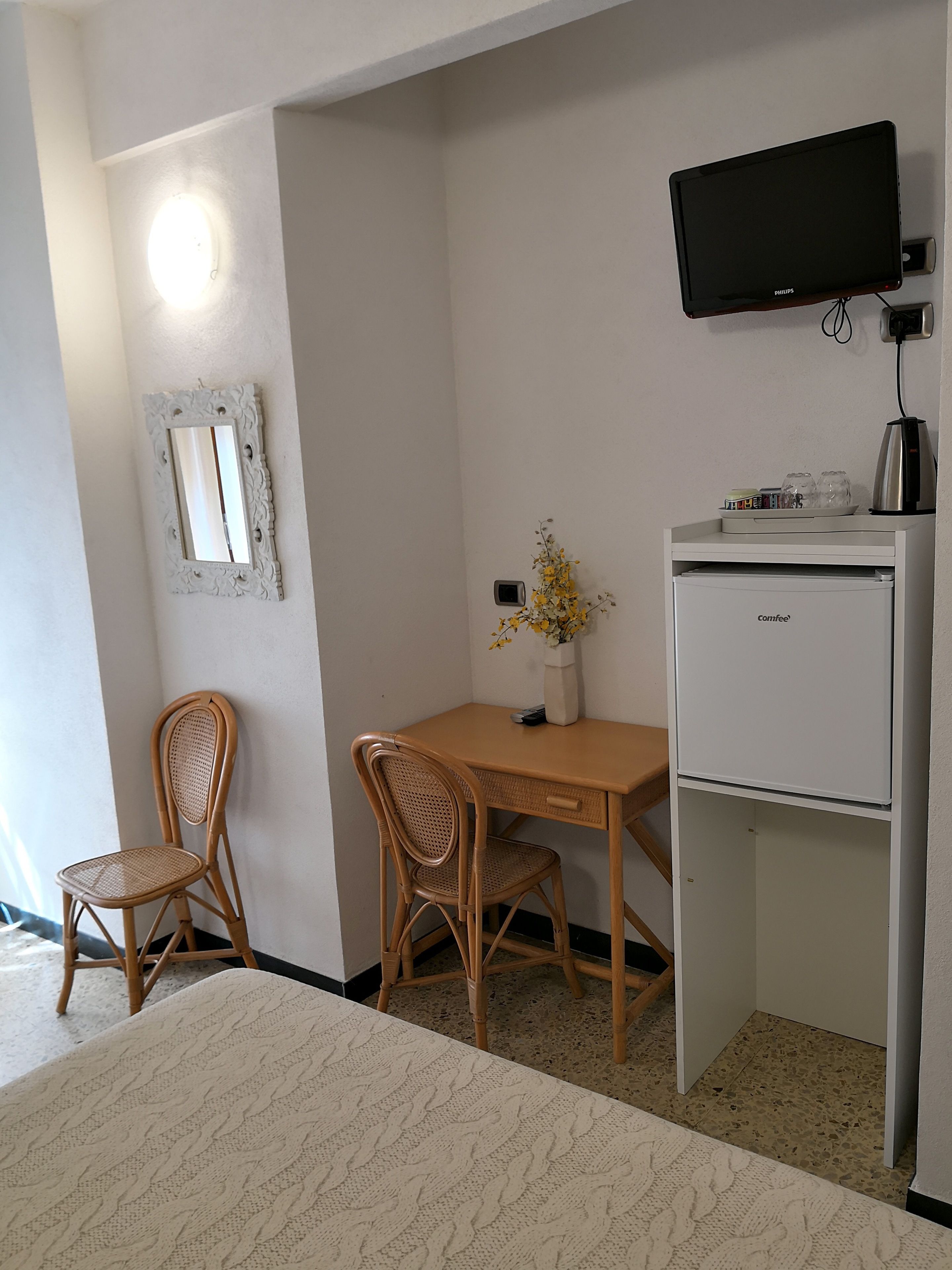 Una habitación pequeña con una cama, un escritorio, dos sillas de mimbre, un televisor, una cafetera y un espejo.