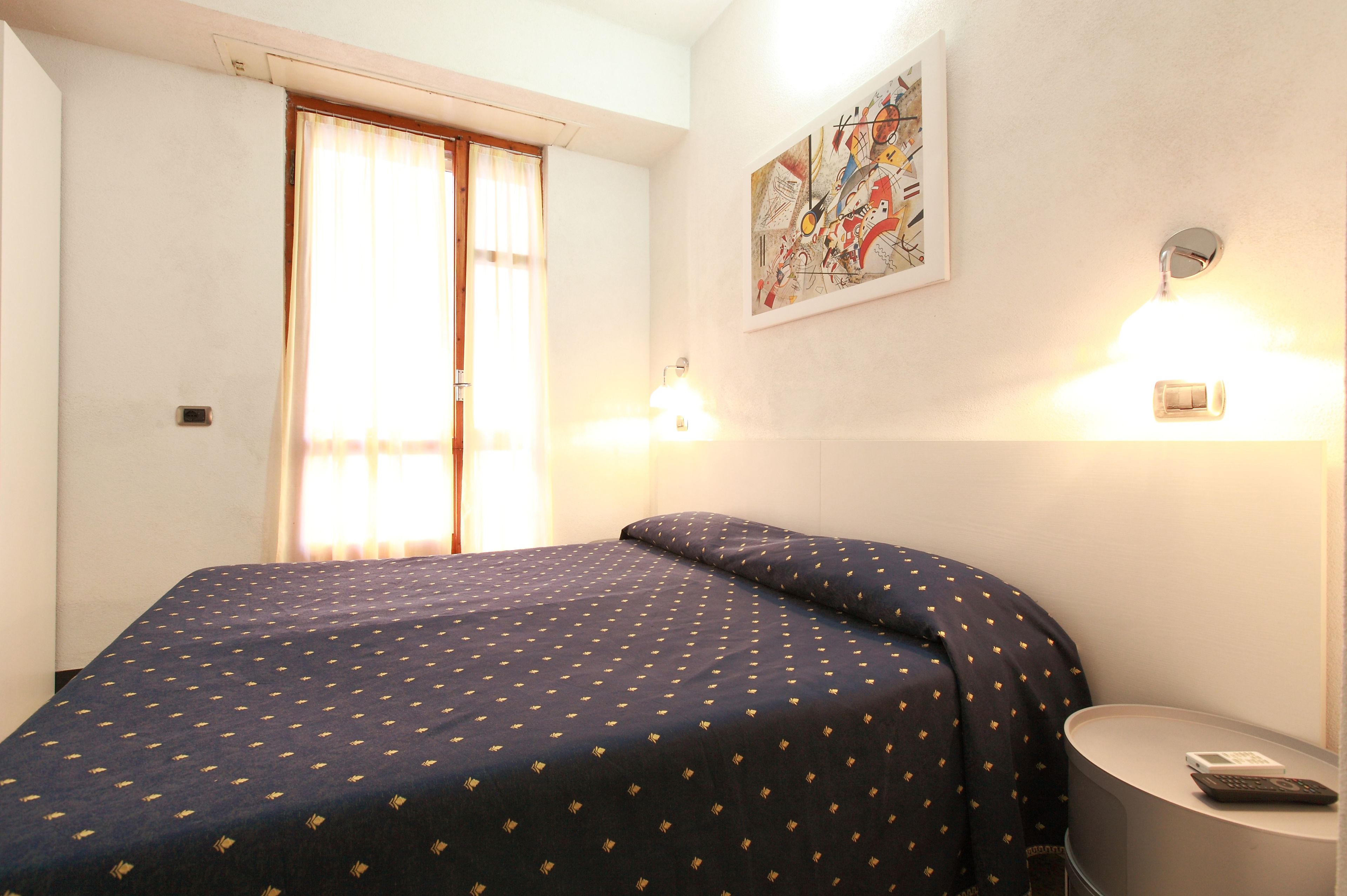 A hotel room with a bed, a nightstand, a lamp, a window with curtains, and a wall art piece.