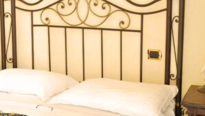 A bed with a metal headboard, a bedside table with a lamp, and a small table with a vase of flowers. - Hotel Sovrano (Alberobello)