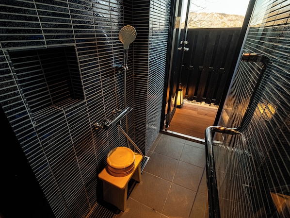 A bathroom with a toilet, a shower with a handheld showerhead, and a window with a view of a wooden deck.