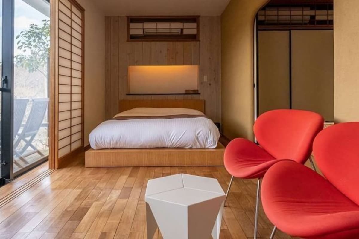 Une chambre avec un plancher en bois, un lit avec un matelas blanc, une chaise rouge et une table de chevet blanche.