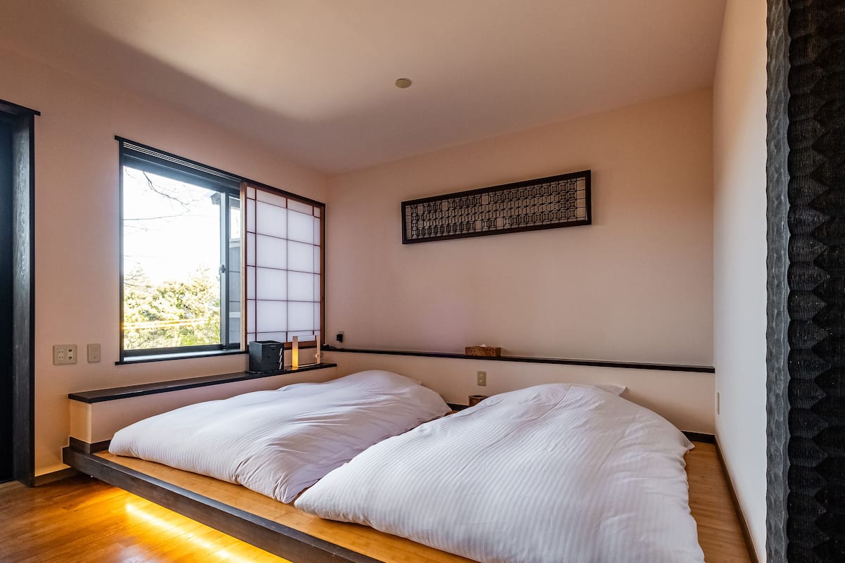 Un dormitorio minimalista con una cama baja tipo plataforma, una ventana con celosía japonesa y una obra de arte mural geométrica.