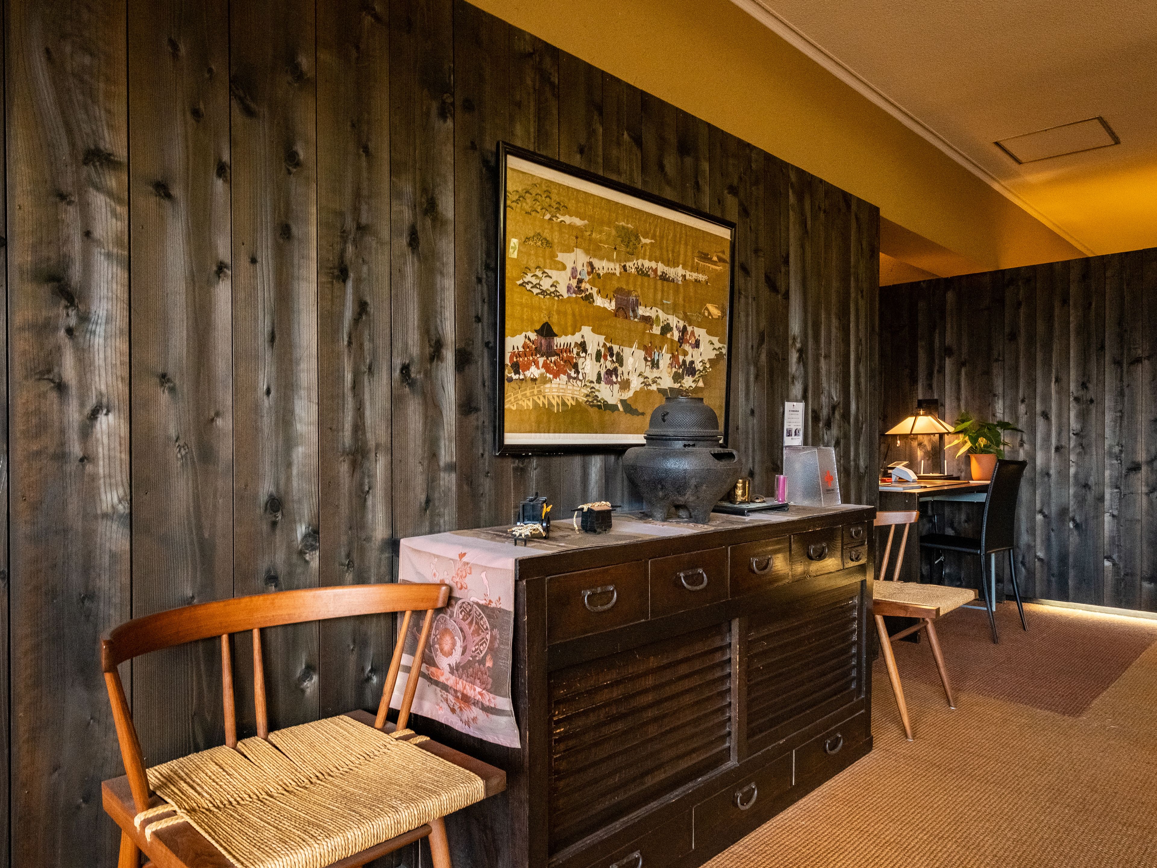 A room with wooden panel walls, a large framed artwork, a traditional stove, and a desk with a lamp and plant.
