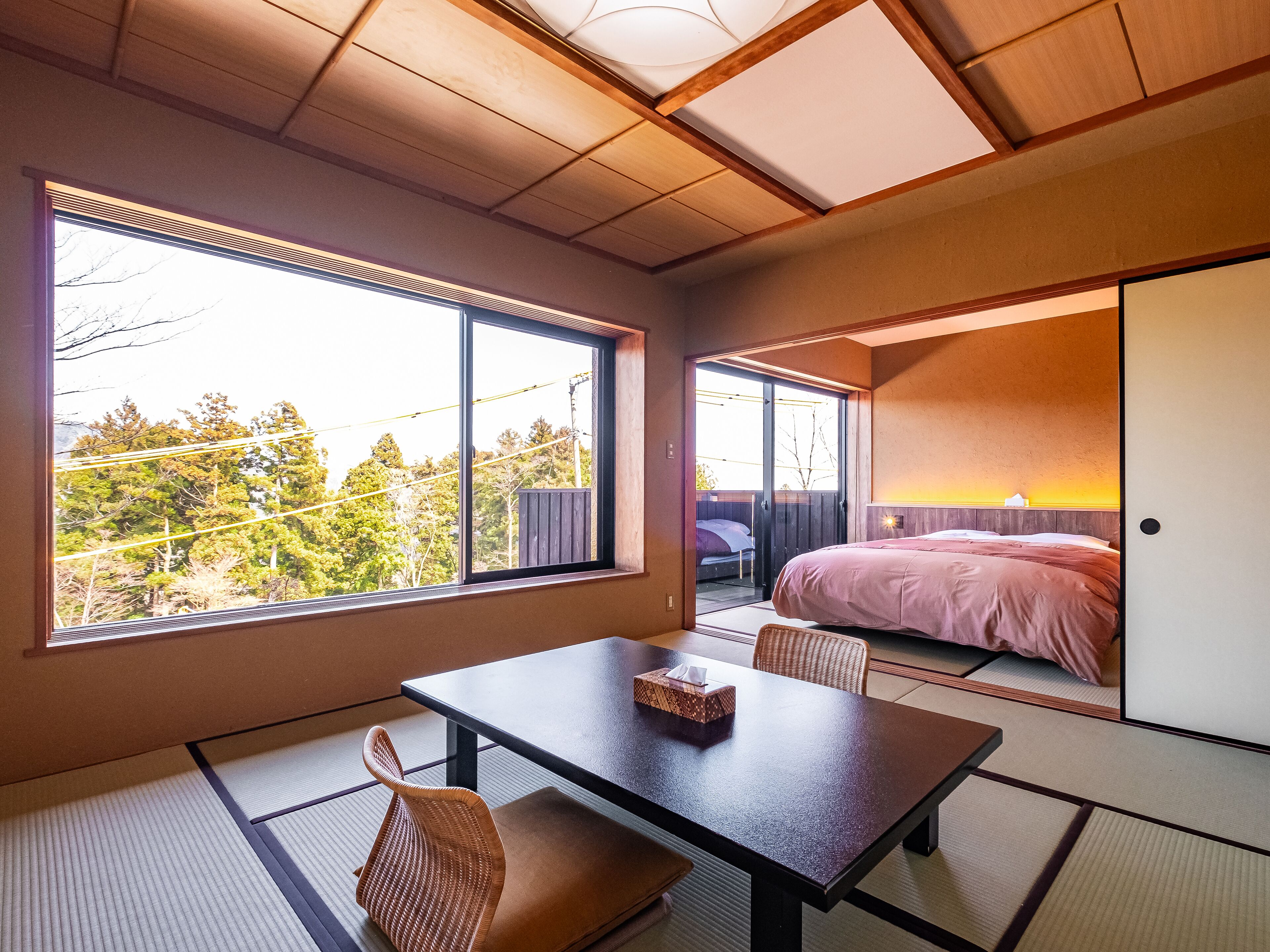 Ein modernes Hotelzimmer mit Tatami-Böden, einem großen Fenster mit Blick auf Bäume, einem Bett mit rosa Bettdecke und einer Essecke mit Tisch und Stühlen.
