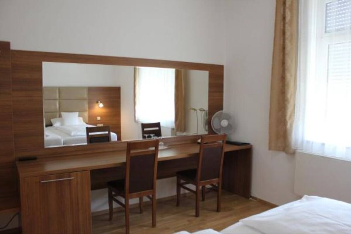 A hotel room with a wooden desk, two chairs, a bed, a large mirror, and a window with curtains.