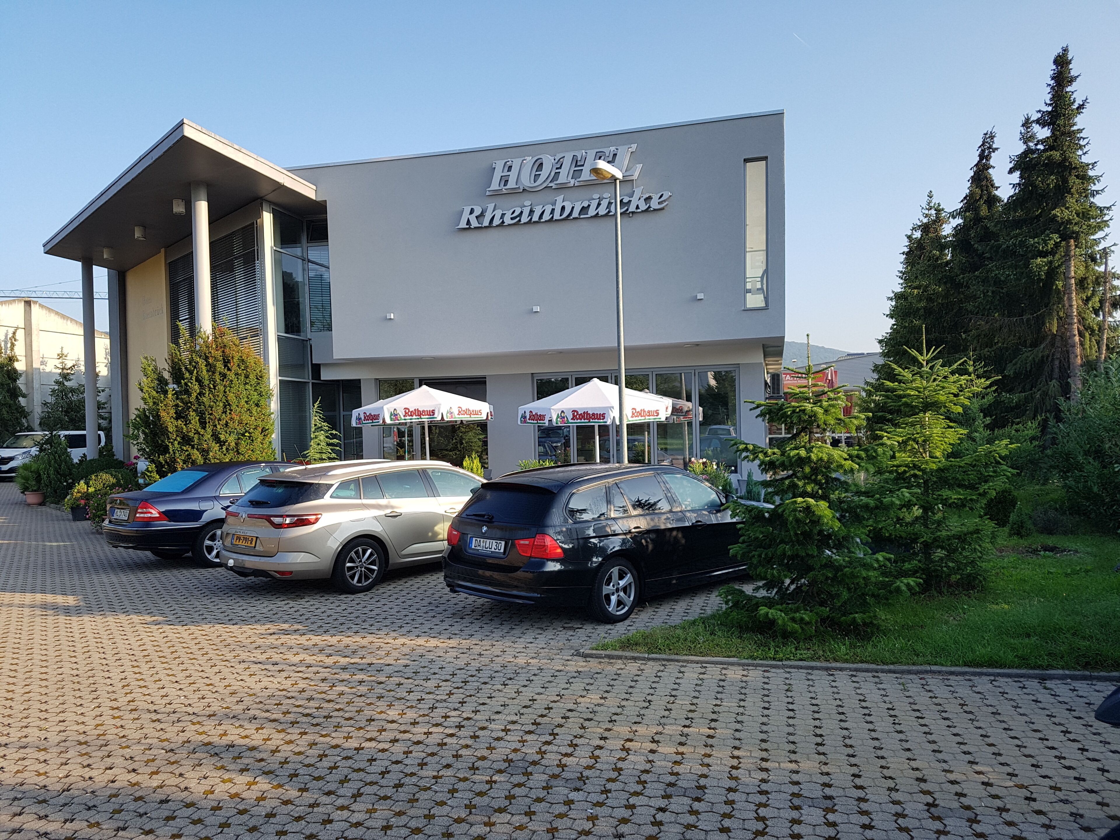 A hotel with a modern design, featuring a large sign with the name "HOTEL Rheinhütte". There are several cars parked in front, and the building is surrounded by greenery and trees.