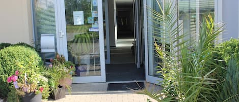 Entrada de un edificio con puerta de cristal, un sendero pavimentado y plantas en macetas.