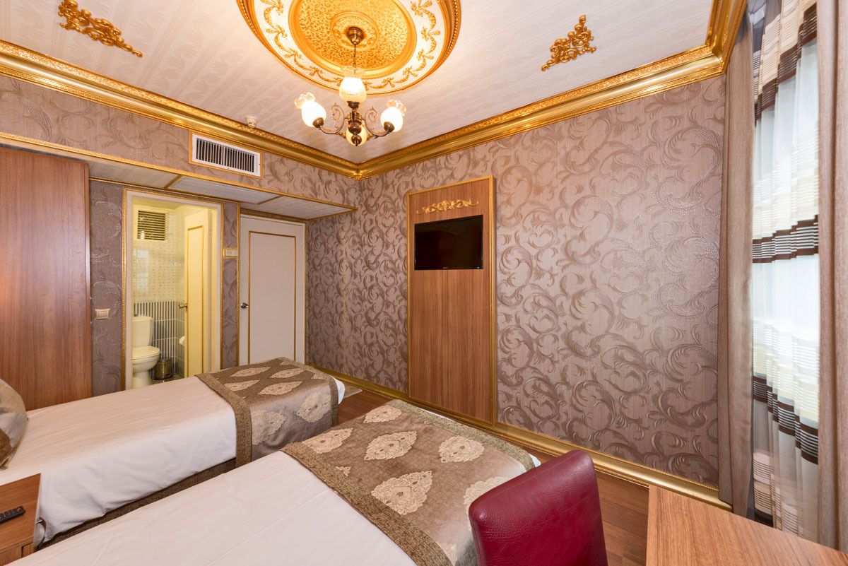 Quarto de hotel com duas camas, televisão, uma cadeira vermelha e teto decorativo.