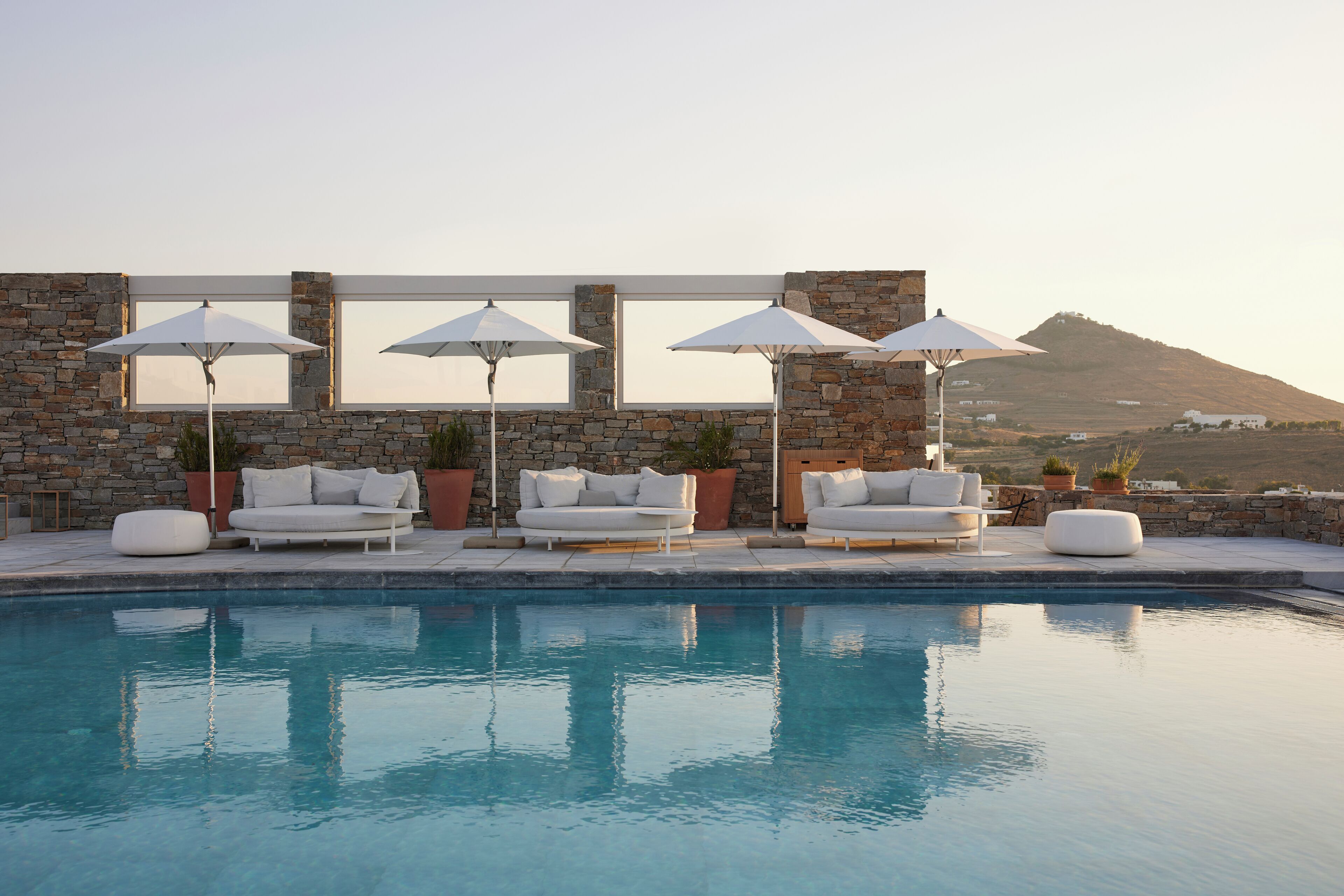 A poolside area with white outdoor furniture and umbrellas, stone walls, and a view of a hill.