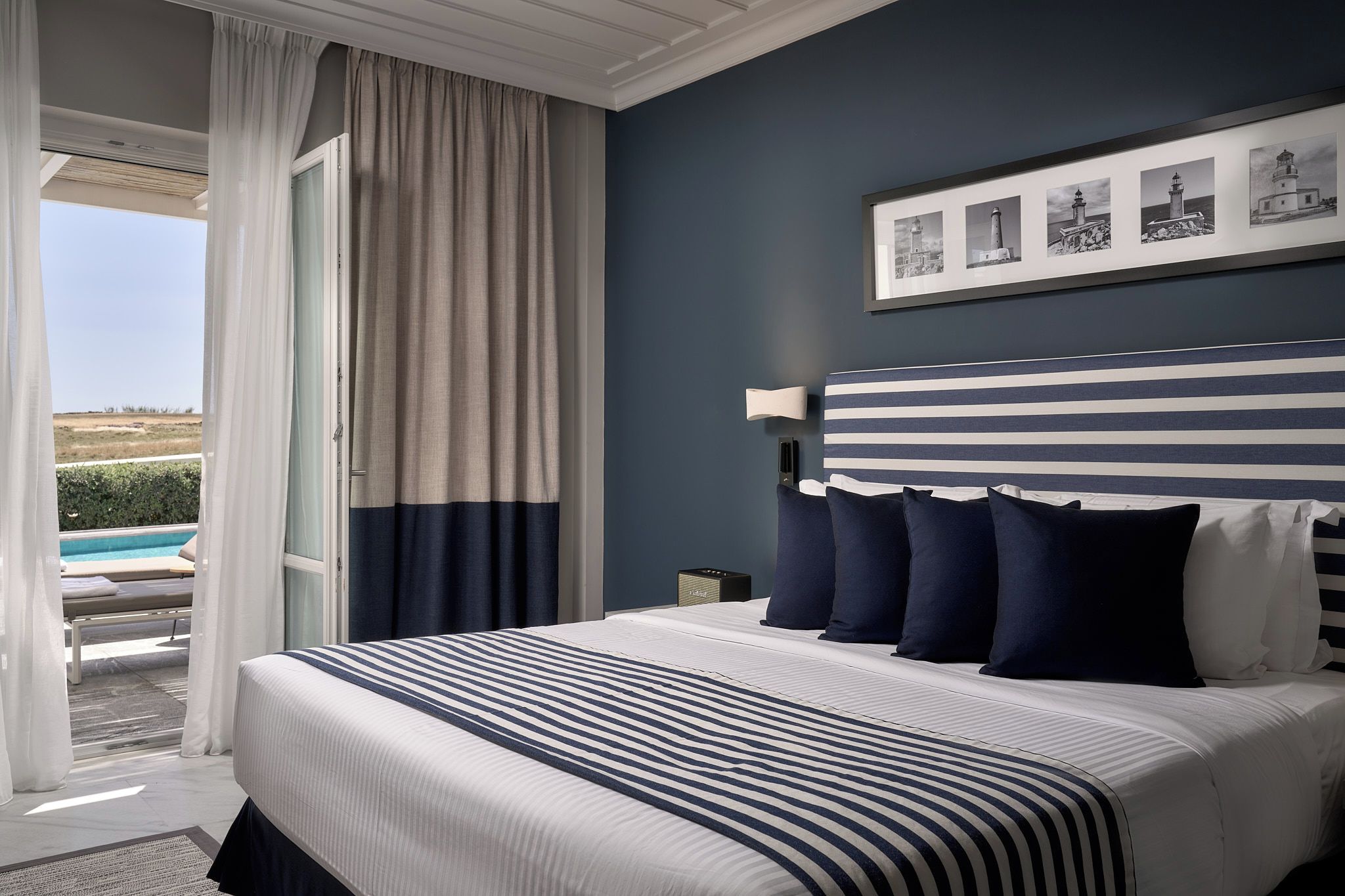 Quarto de hotel com uma cama grande, vista para uma piscina e um quadro com faróis na parede.